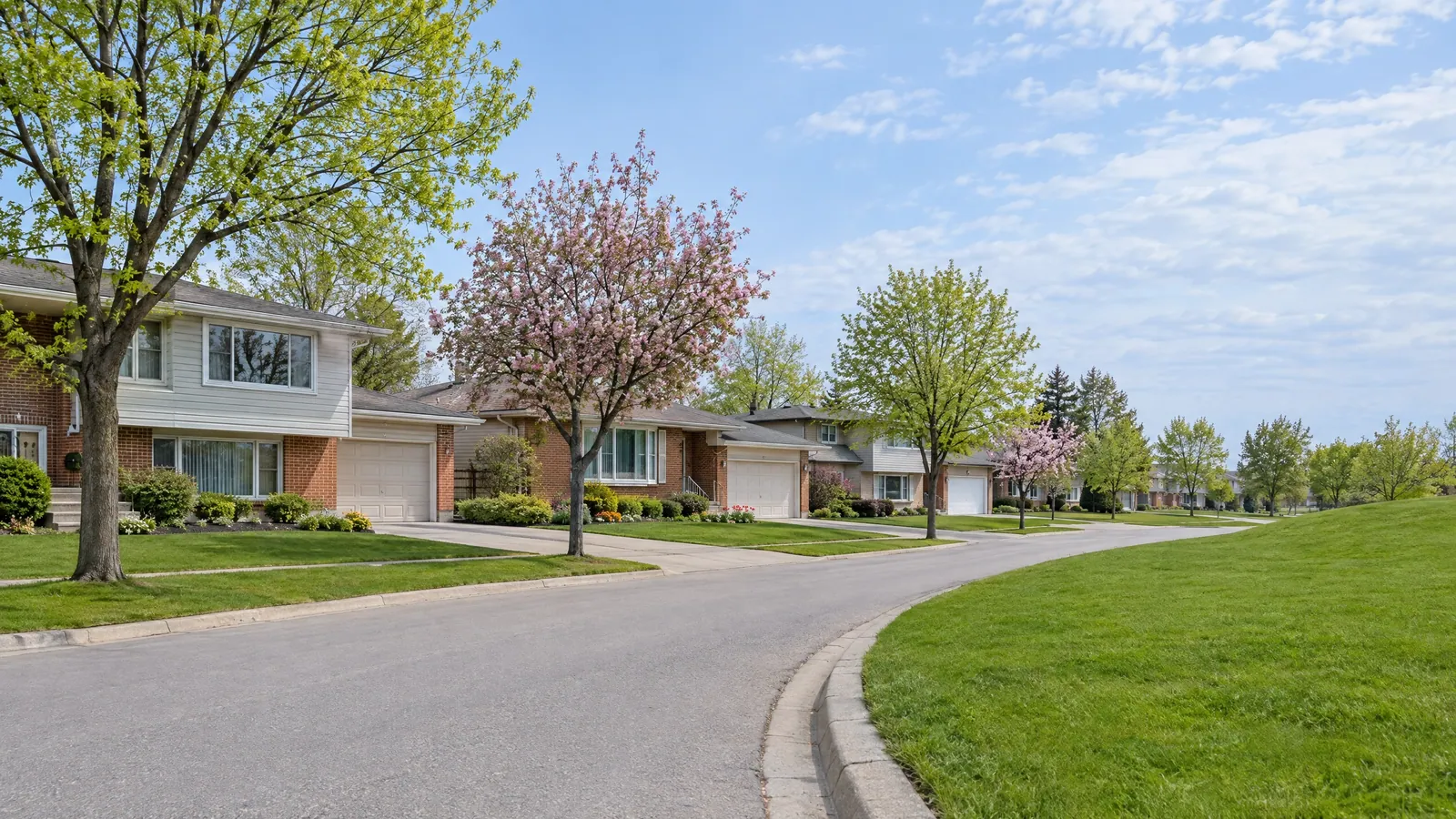 Curving spring street of 1970s back-split houses in brick and aluminium siding, with slider windows and wide picture windows
