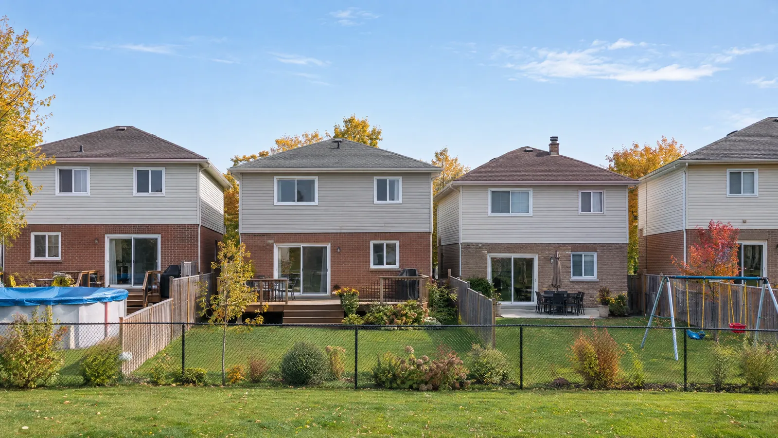 Backyards of two-storey brick-and-siding homes, each with a glass slider opening onto a deck or patio, seen across a chain-link fence in autumn
