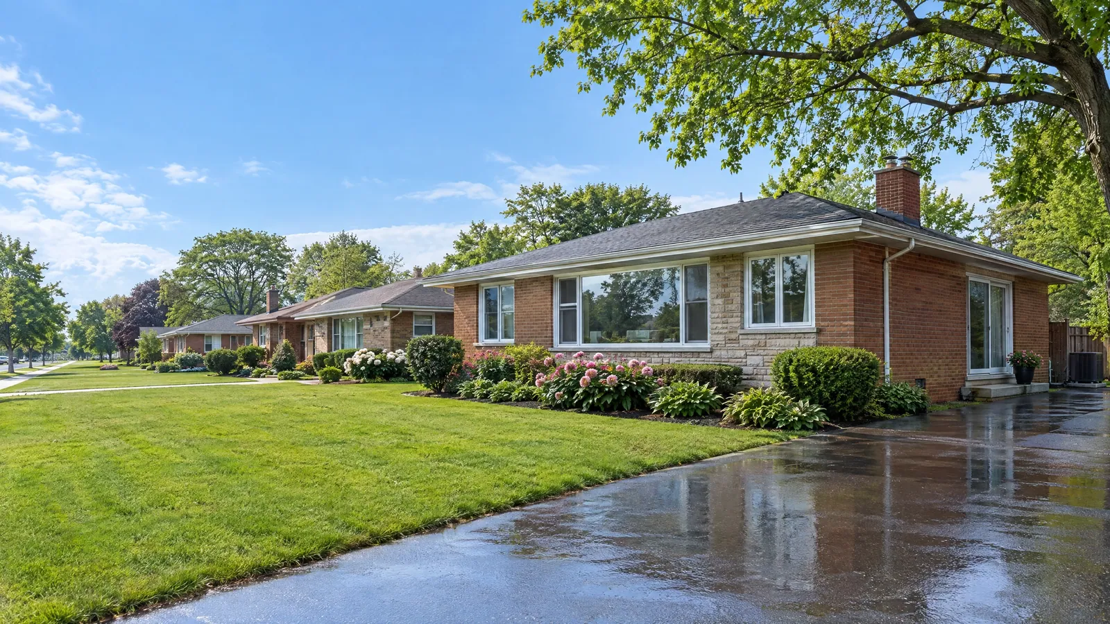 Brick ranch bungalow with a large picture window and a sliding patio door at the side, next to a rain-wet driveway and a row of similar homes